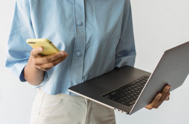 A woman standing with a laptop while holding a mobile phone, preparing to make a call or communicate.